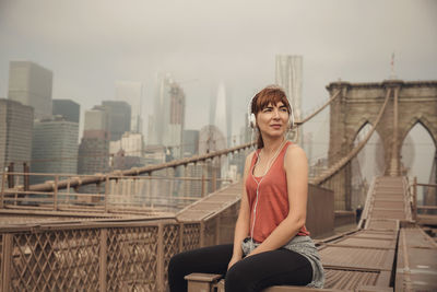 Woman listening to music while sitting on bridge against sky