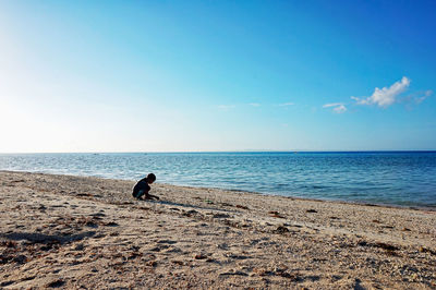 Side view of boy crouching on shore at beach against sky