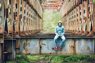 Full length of young man using phone while sitting on abandoned bridge