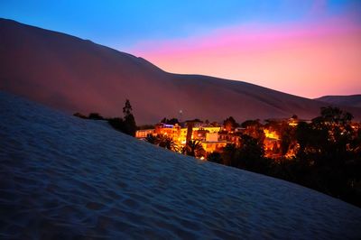 Illuminated buildings by silhouette mountain against sky at sunset