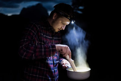 Young man preparing food using headlamp in forest at night