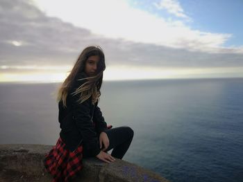 Portrait of girl sitting on cliff by sea against cloudy sky