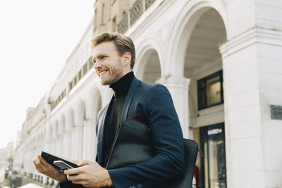 Smiling businessman with smart phone looking away while standing against building in city