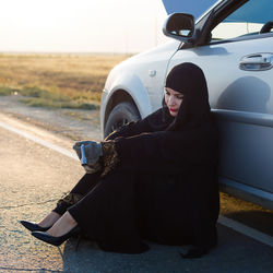 Woman wearing hijab sitting by damaged car on road