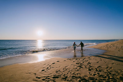 Couple walking at beach against clear sky during sunset