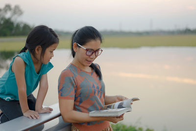 Mother reading book to daughter by lake during sunset