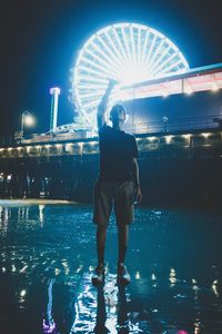 Rear view of woman standing in illuminated amusement park against sky at night