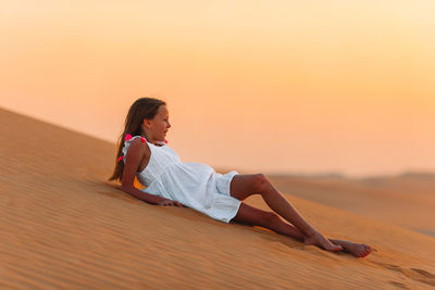 Side view of woman sitting on beach against sky during sunset