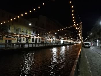 Illuminated street by river against buildings at night