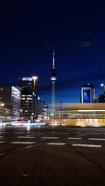 Light trails on city street by buildings against sky at night