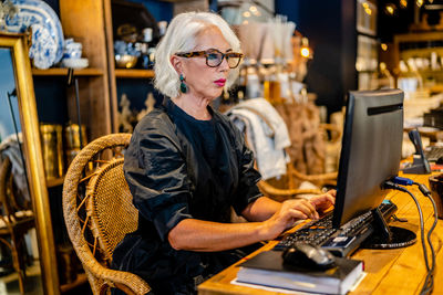 Side view of mature female entrepreneur in elegant dress sitting at table and typing on keyboard while working on computer on project online