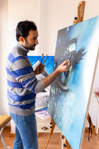 Side view of young bearded ethnic painter in casual wear painting with brush on canvas in art studio