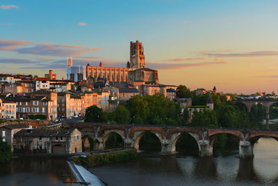 Bridge over river by buildings against sky at sunset