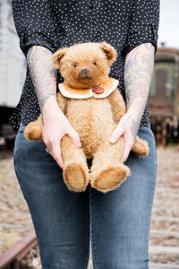 A young caucasian girl with glasses and tattoos smiles, a brown teddy bear