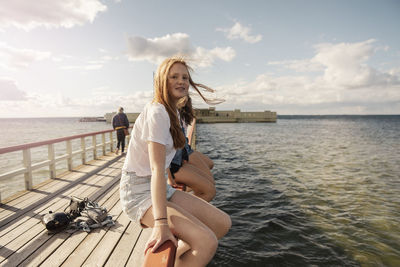 Female friends sitting over railing amidst sea against sky