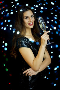 Portrait of beautiful smiling young woman drinking champagne against illuminated lights
