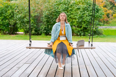 Happy woman swinging on swing in city park, wearing yellow dress and blue coat