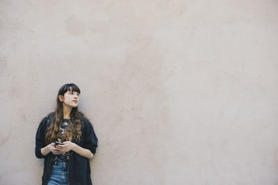 Thoughtful female computer programmer holding smart phone while standing against beige wall