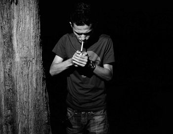 Young man lighting cigarette while standing by tree
