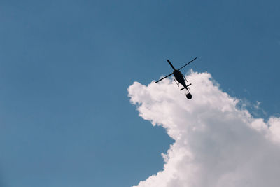 Silhouette of a firefighter helicopter against a background of white clouds and blue sky.