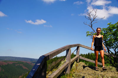 Low angle portrait of young woman standing on mountain against blue sky during sunny day