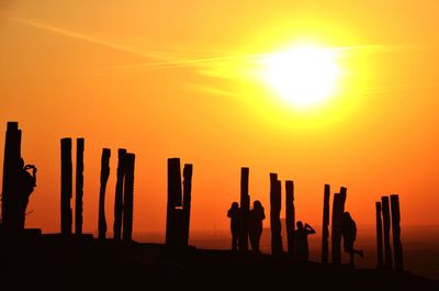 Silhouette people standing amidst wooden posts against sky during sunrise