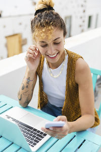 Happy young woman surfing net through mobile phone sitting at table on rooftop