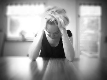 Depressed woman with hand in hair sitting at wooden table in brightly lit room