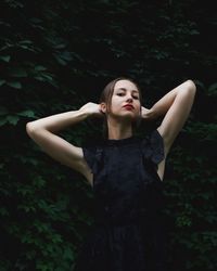 Portrait of beautiful young woman with hands behind head standing against plants