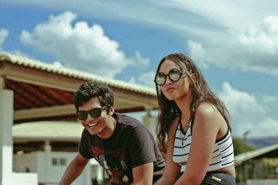 Portrait of friends wearing sunglasses sitting against cloudy sky