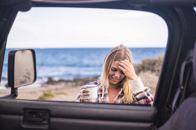 Young woman holding coffee cup at beach seen through car window