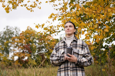 A woman in warm clothes in the autumn forest holds a glass with a hot drink and smiles