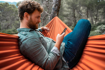 Man sitting using smart phone while on hammock in forest