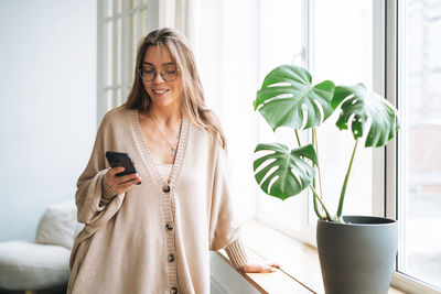 Young beautiful woman with in cozy knitted cardigan using mobile phone in bright interior at home