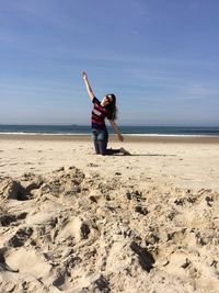 Full length of young woman kneeling on sand at beach against sky