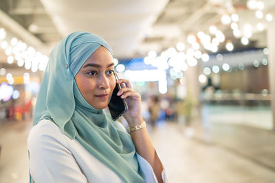 Young woman wearing hijab talking on smart phone while standing in restaurant