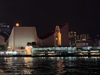 Illuminated buildings by river against sky at night