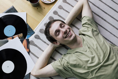 High angle view of happy man lying down near vinyl records on carpet at home