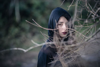 Portrait of young woman wearing hooded shirt standing by bare tree in forest