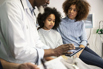 Woman by ill daughter looking at doctor sharing report on tablet pc at hospital