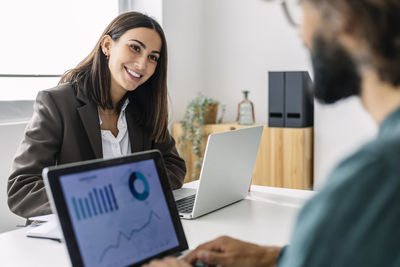 Smiling young businesswoman with laptop looking at colleague in office