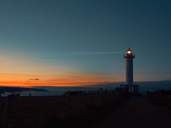 Silhouette lighthouse by building against sky during sunset