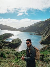 Portrait of young man standing against lake amidst mountains against sky