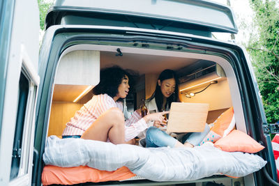 Young multiracial female friends drinking beer and using laptop together while preparing content for travel blog inside camper van during summer adventure in nature