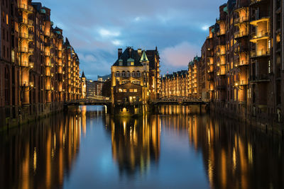 Illuminated buildings by river against cloudy sky at dusk
