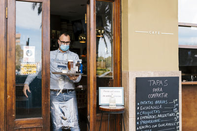 Waiter wearing face mask holding drinks in tray for customers outside a bar