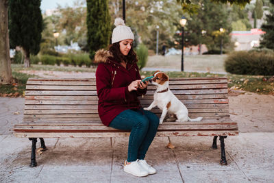 Woman with dog using phone while sitting on bench against trees