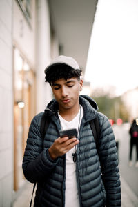 Teenage boy wearing cap using smart phone while walking on footpath in city