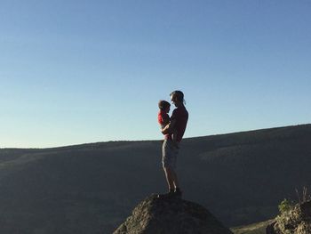 Man carrying son on rock against clear blue sky