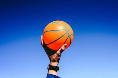 Cropped hand holding basketball against blue sky